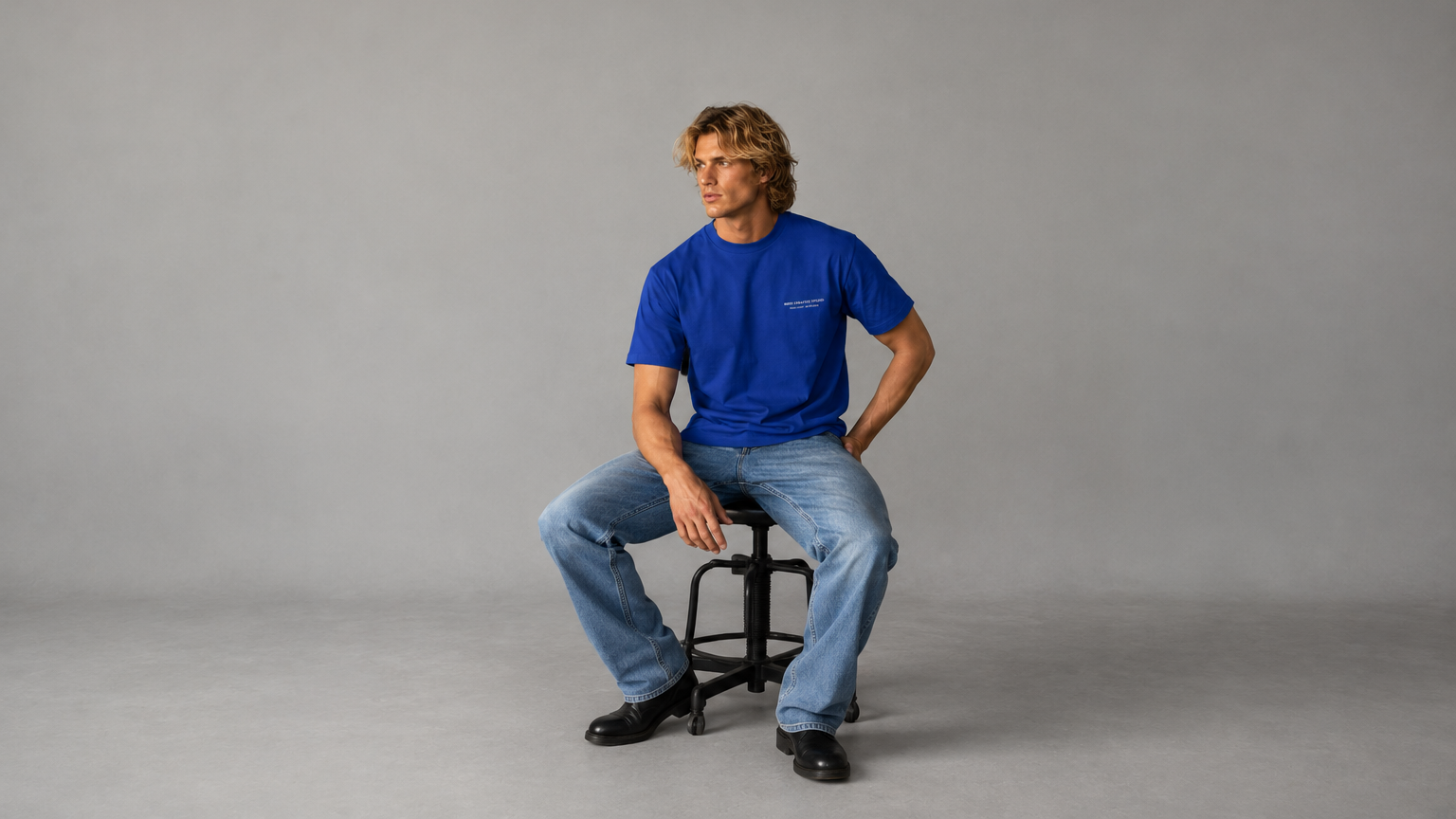 Blonde male model posing whilst sitting on a stool wearing a the  "Meek Creative Studios" T shirt in royal blue